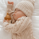 Baby wearing a cream knitted hat and jumper asleep holding with a Hevea Glass Baby Bottle