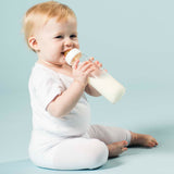 Baby sitting on the floor drinking milk from Hevea Glass Baby Bottles Standard Neck - 240ml.
