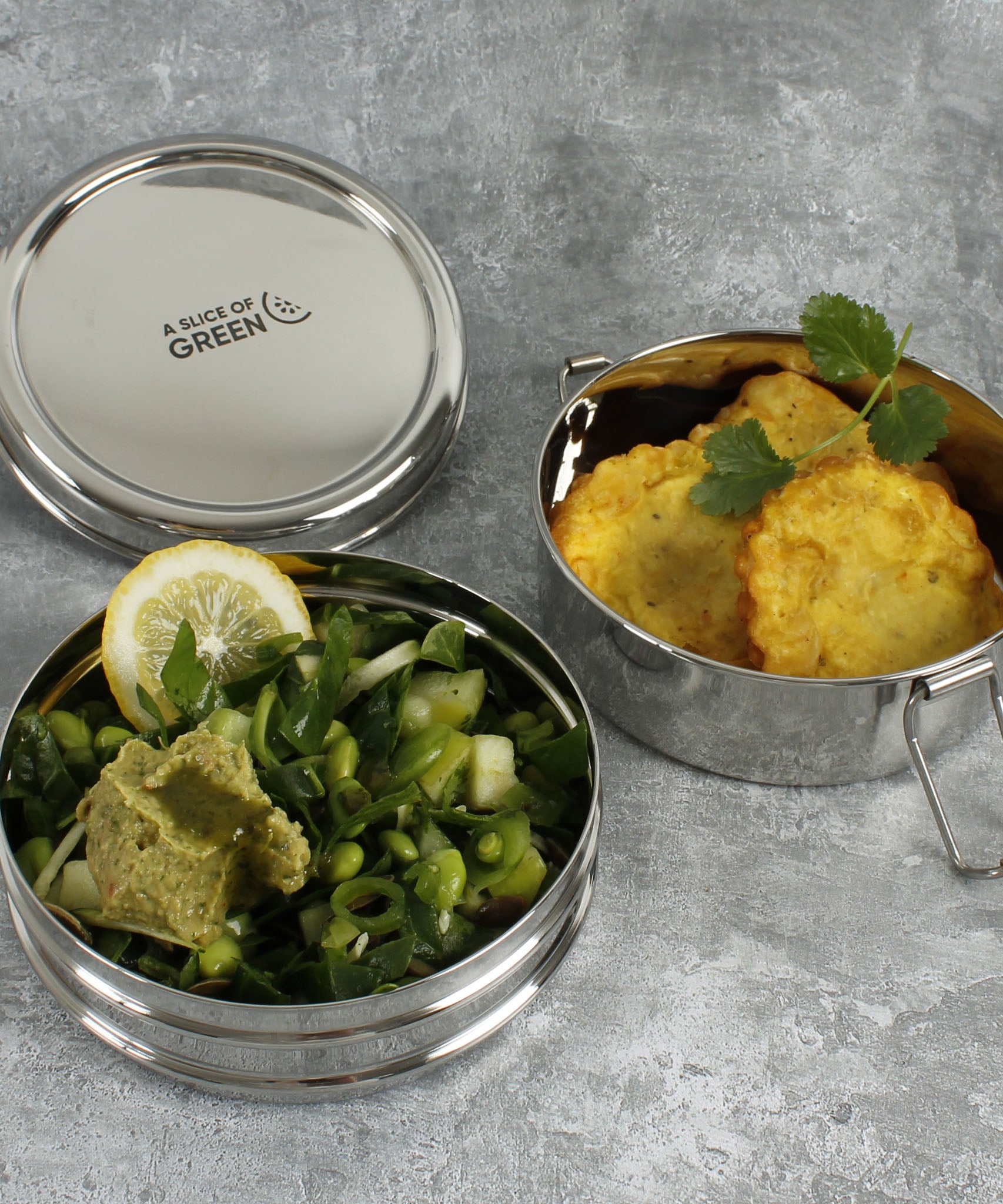 A close up of various food items inside 2 of the tins from the stainless steel A slice of green chapra two tier round lunchbox.