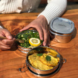 An adult eating food from the stainless steel A slice of green chapra two tier round lunchbox.