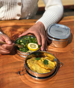 An adult eating food from the stainless steel A slice of green chapra two tier round lunchbox.