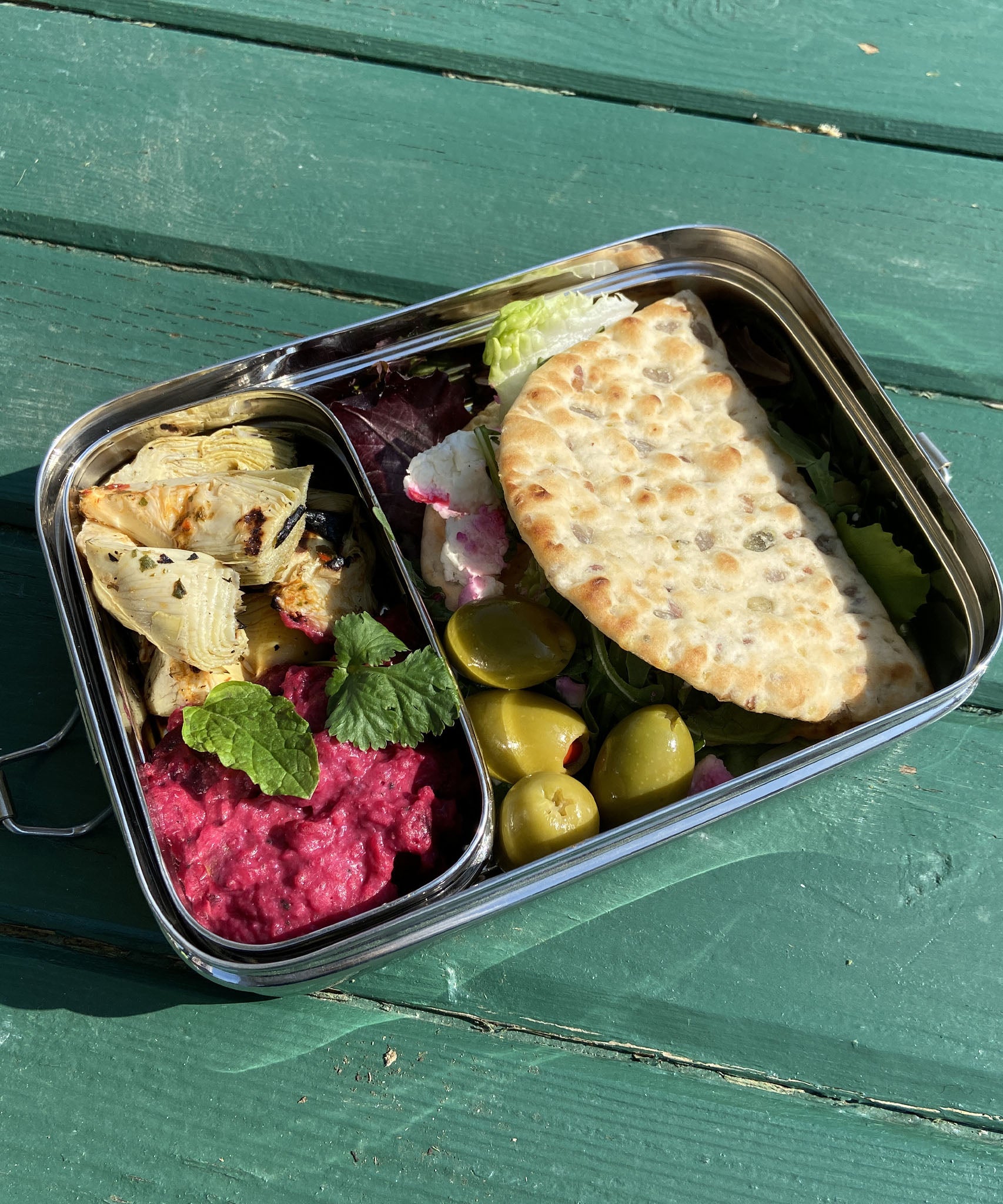 A close up of the A slice of green large lunchbox with mini container on a wooden table with various food items inside