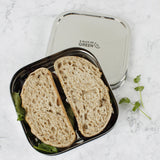 A top down image of A slice of green shimla large square container lunchbox resting on top of the lid with a sandwich inside