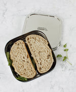 A top down image of A slice of green shimla large square container lunchbox resting on top of the lid with a sandwich inside
