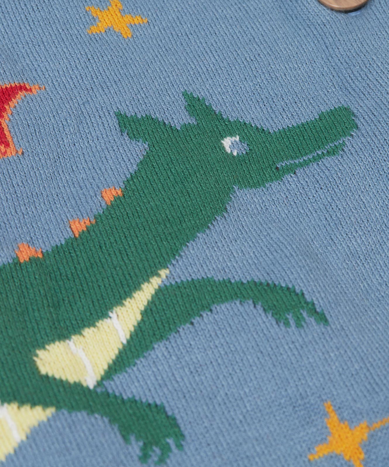 A close up of the green dragon on the front of the Frugi Organic Cotton Reva Dragon Skies Knitted Kids' Cardigan. This blue cardigan features a fun, colourful design featuring a green dragon, stars, rainbow, clouds and a mushroom. This cardigan is a part of a wide range of organic cotton clothing for children available here at Babipur.