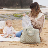 An adult and child sat on the beach, with the child playing with toys and the adult holding a white and blue cloth with the Sage nappy bag in front of them