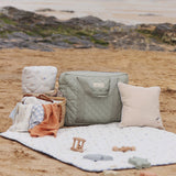The Avery Row Nappy Changing Bag - Sage placed on the beach with other Avert Row products next to it