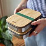 An adult holding a pair of Black + Blum airtight lunchboxes in olive available at Babipur. Lunchboxes feature a Bamboo lid with a olive green silicone band holding the lid and lunchbox.