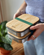 An adult holding a pair of Black + Blum airtight lunchboxes in olive available at Babipur. Lunchboxes feature a Bamboo lid with a olive green silicone band holding the lid and lunchbox.