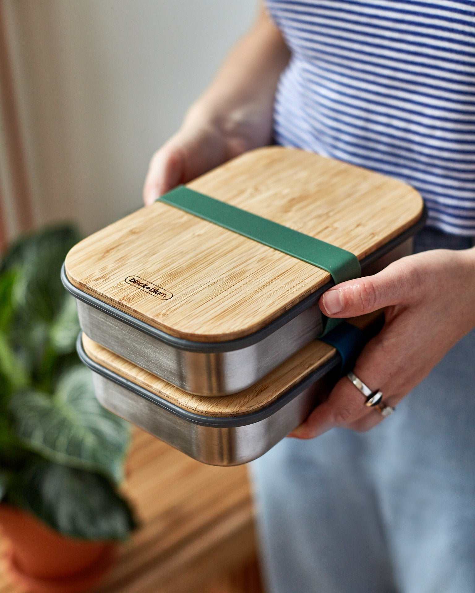 An adult holding a pair of Black + Blum airtight lunchboxes in olive available at Babipur. Lunchboxes feature a Bamboo lid with a olive green silicone band holding the lid and lunchbox.