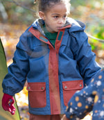 Girl in a forest wearing a hooded Little Green Radicals Waterproof Winter School Coat, made from recycled bottles, PFC Free, with a Moonlight Blue print with deep red details down the zip and front pockets, for sale at Babipur.