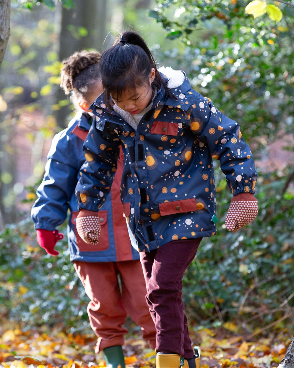 Child playing in a forest wearing a Kids Warm Waterproof Winter School Coat in a navy night moon print, blue and floral, by Little Green Radicals, made from recycled bottles and a cosy polyester fleece, buy now at Babipur.