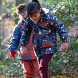 Child playing in a forest wearing a Kids Warm Waterproof Winter School Coat in a navy night moon print, blue and floral, by Little Green Radicals, made from recycled bottles and a cosy polyester fleece, buy now at Babipur.