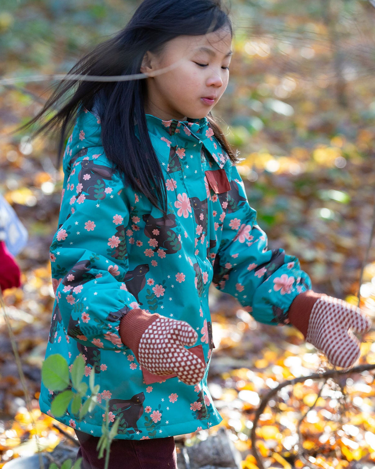 Child playing in a forest wearing a Kids Warm Waterproof Winter School Coat in Winter Rabbits print, blue and floral, by Little Green Radicals, made from recycled bottles and a cosy polyester fleece, buy now at Babipur.