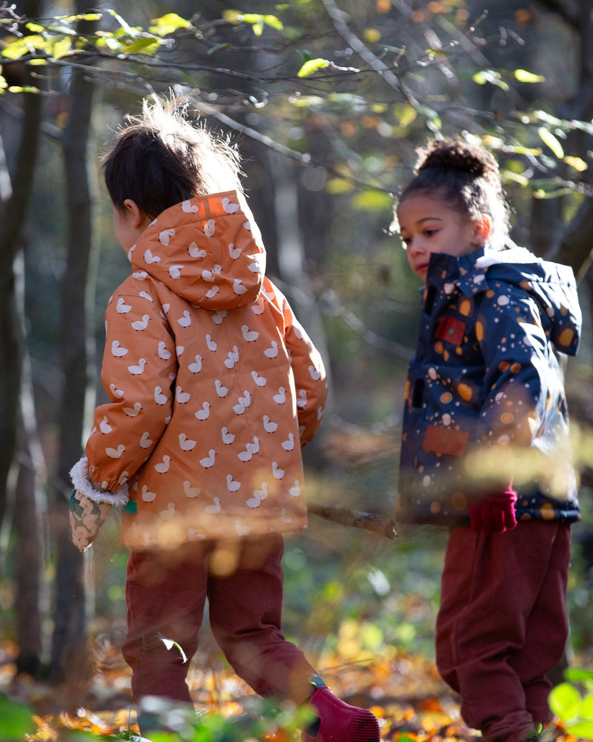 Back of boy playing in a forest wearing a hooded Little Green Radicals Waterproof Winter School Coat, made from recycled bottles, with a yellow gold duck print, for sale at Babipur.