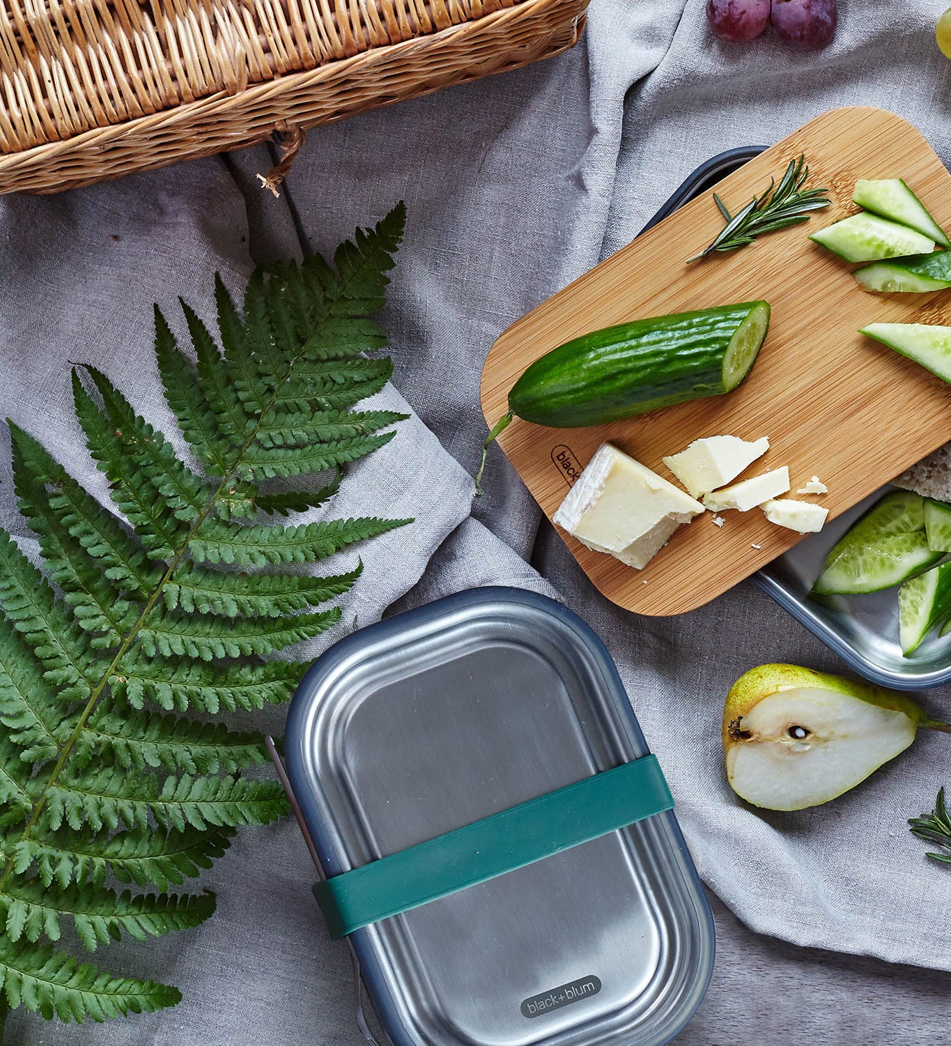 Black + Blum leakproof stainless steel lunchbox with a olive silicone holder on a grey blanket surrounded by different foods with a bamboo lunchbox lid used to prep food