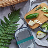 Black + Blum leakproof stainless steel lunchbox with a olive silicone holder on a grey blanket surrounded by different foods with a bamboo lunchbox lid used to prep food