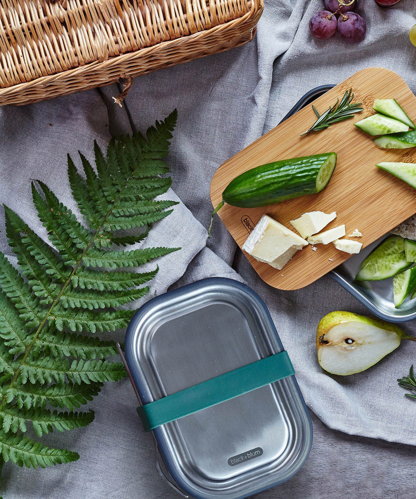 Black + Blum leakproof stainless steel lunchbox with a olive silicone holder on a grey blanket surrounded by different foods with a bamboo lunchbox lid used to prep food