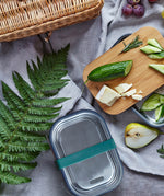 Black + Blum leakproof stainless steel lunchbox with a olive silicone holder on a grey blanket surrounded by different foods with a bamboo lunchbox lid used to prep food