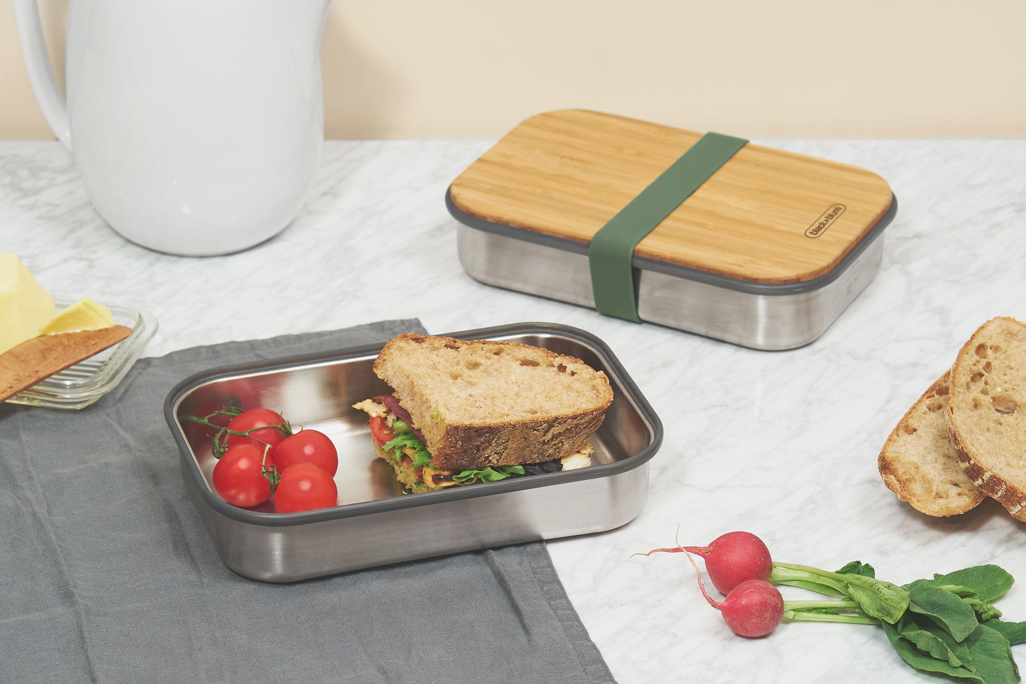 A sandwich and tomatoes inside a Black + Blum stainless steel airtight stainless steel sandwich box with a olive silicone holder available at Babipur.