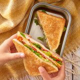 A close up of a adult hand holding a toasted sandwich with another in a Black + Blum stainless steel airtight stainless steel sandwich box