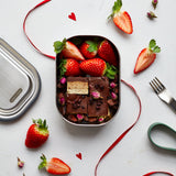 A top down view of the Black + Blum leakproof stainless steel lunchbox with a olive silicone holder on top of red decorative ribbon with a chocolatey desert inside the lunchbox