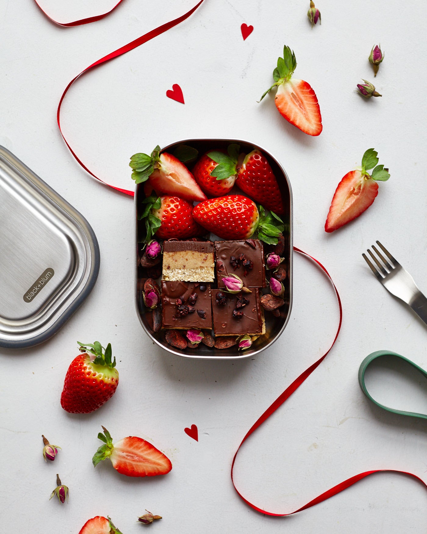 A top down view of the Black + Blum leakproof stainless steel lunchbox with a olive silicone holder on top of red decorative ribbon with a chocolatey desert inside the lunchbox