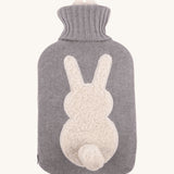 The Ecoliving Natural Rubber Hot Water Bottle - Bunny, has a soft grey knitted cotton cover with a fluffy white bunny silhouette and a cute white cotton tail. The image is on a cream background