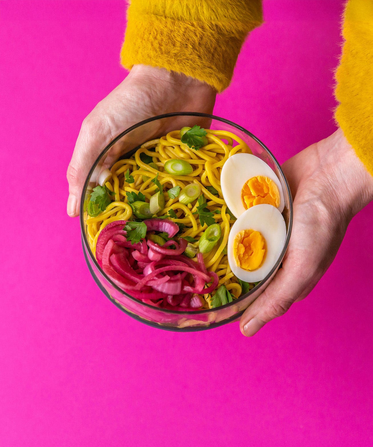 An adult hand holding the Elephant box large lunch bowl full of food and against a bright pink background. Lunch bowl available at Babipur.