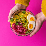 An adult hand holding the Elephant box large lunch bowl full of food and against a bright pink background. Lunch bowl available at Babipur.