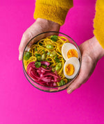 An adult hand holding the Elephant box large lunch bowl full of food and against a bright pink background. Lunch bowl available at Babipur.