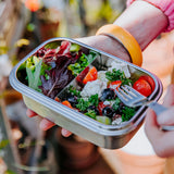 A close up of the Elephant Box Clip & Seal Lunchbox With Removable Divider - 800ml with a salad inside. A person is holding the lunch box in one hand, and a Elephant Box Spork in the other