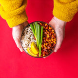 An adult hand holding the Elephant box large lunch bowl full of food and against a bright red background. Lunch bowl available at Babipur.