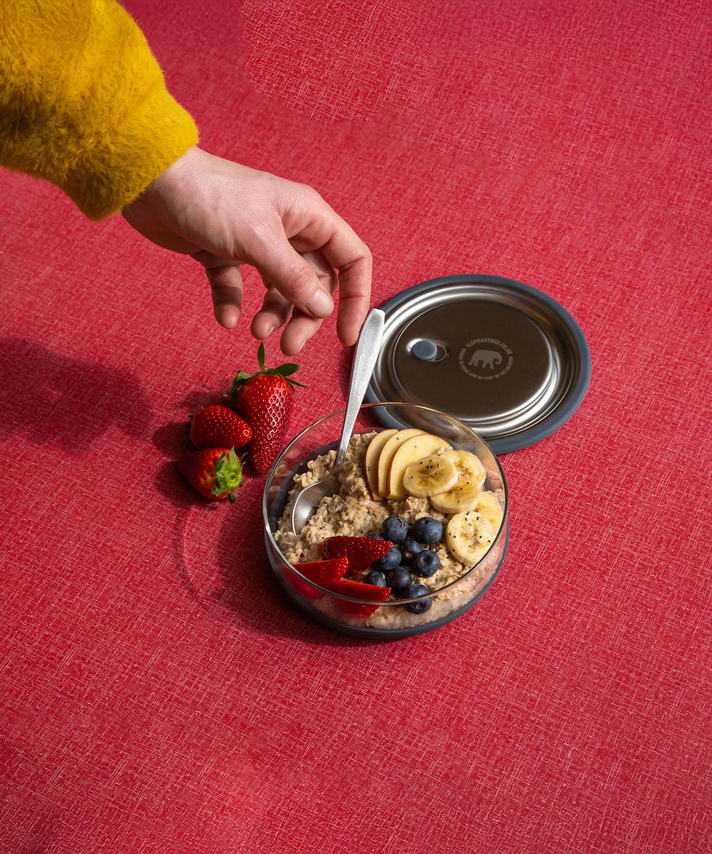 An adult hand reaching out for a spoon in the Elephant box medium lunchbowl full of different fruit. Lunch bowl available at Babipur