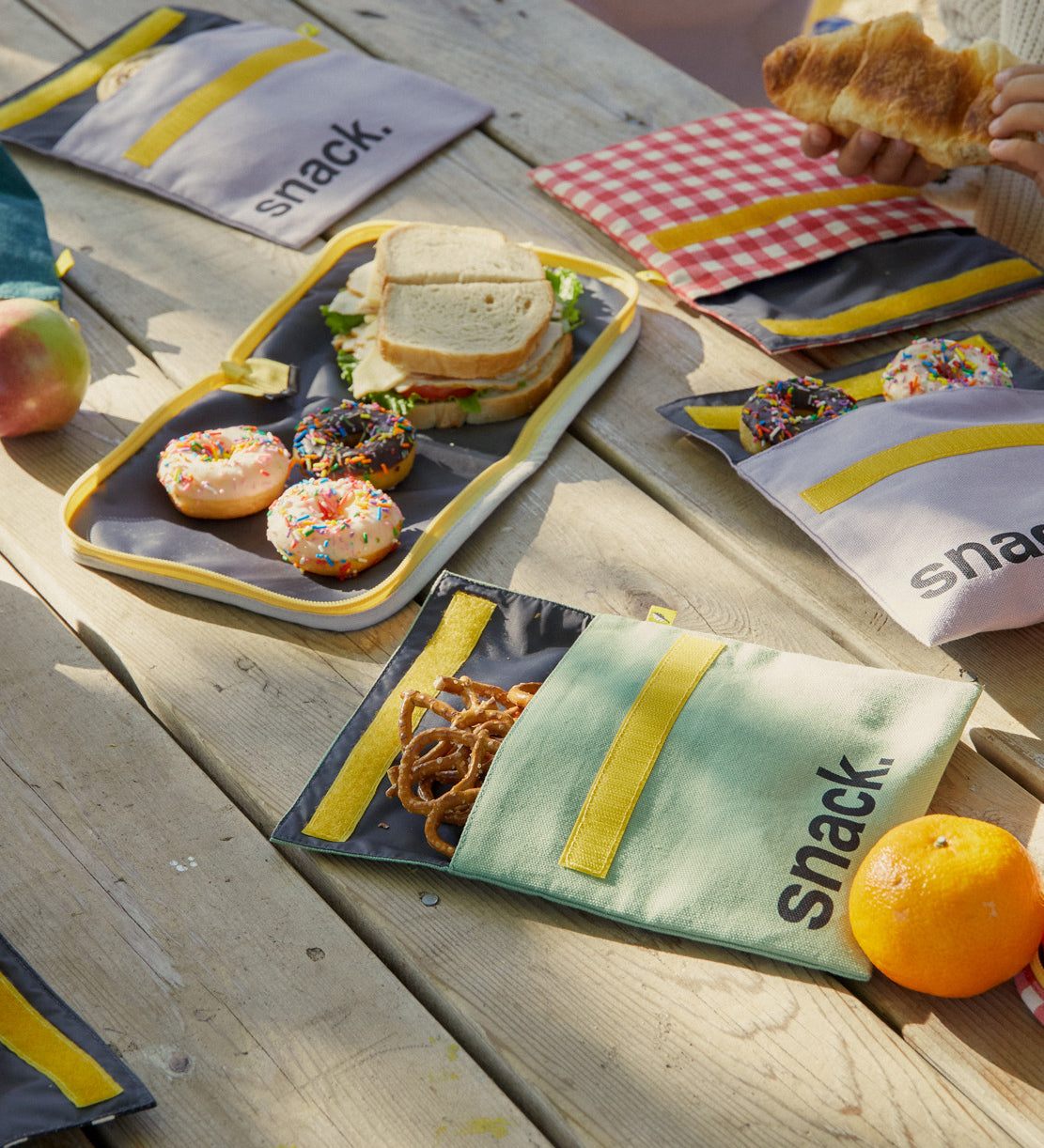 A selection of Fluf Flip Organic Cotton Snack Bags filled with snack on a picnic table outdoors. Reusable cotton bags with the word 'snack written in black front on the front.