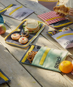 A selection of Fluf Flip Organic Cotton Snack Bags filled with snack on a picnic table outdoors. Reusable cotton bags with the word 'snack written in black front on the front.