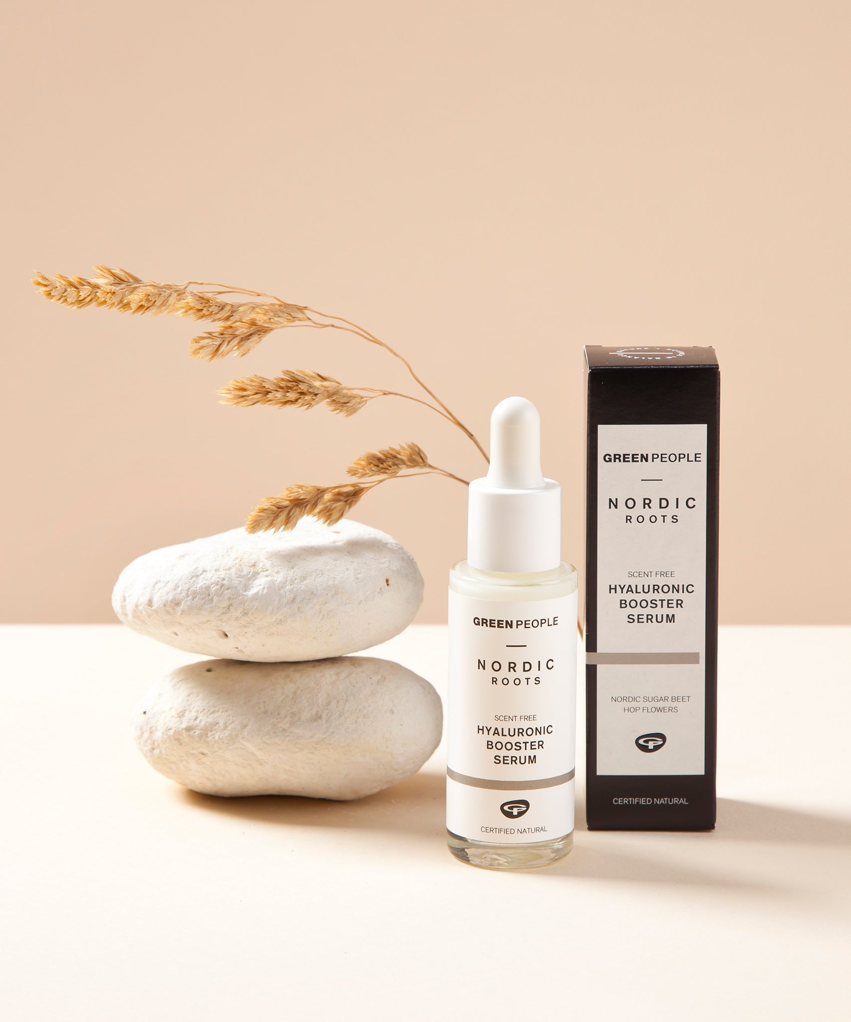 The Green People Nordic Roots Hyaluronic Booster Serum stood next to its box with white rocks just behind the bottle and dried grass