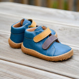 Froddo barefoot first step baby shoe in dark denim available at Babipur standing on a wooden surface. A dark blue natural leather shoe with a 2 velcro fastener in dark yellow and brown for a secure fit. Features a light brown grippy sole