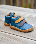 Froddo barefoot first step baby shoe in dark denim available at Babipur standing on a wooden surface. A dark blue natural leather shoe with a 2 velcro fastener in dark yellow and brown for a secure fit. Features a light brown grippy sole