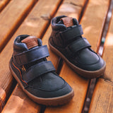 Froddo barefoot tex kids autumn shoes in navy standing on a wooden surface and available at Babipur. Soft ankle high shoes in navy with a light brown rubber sole. Features two velcro straps for a secure fit