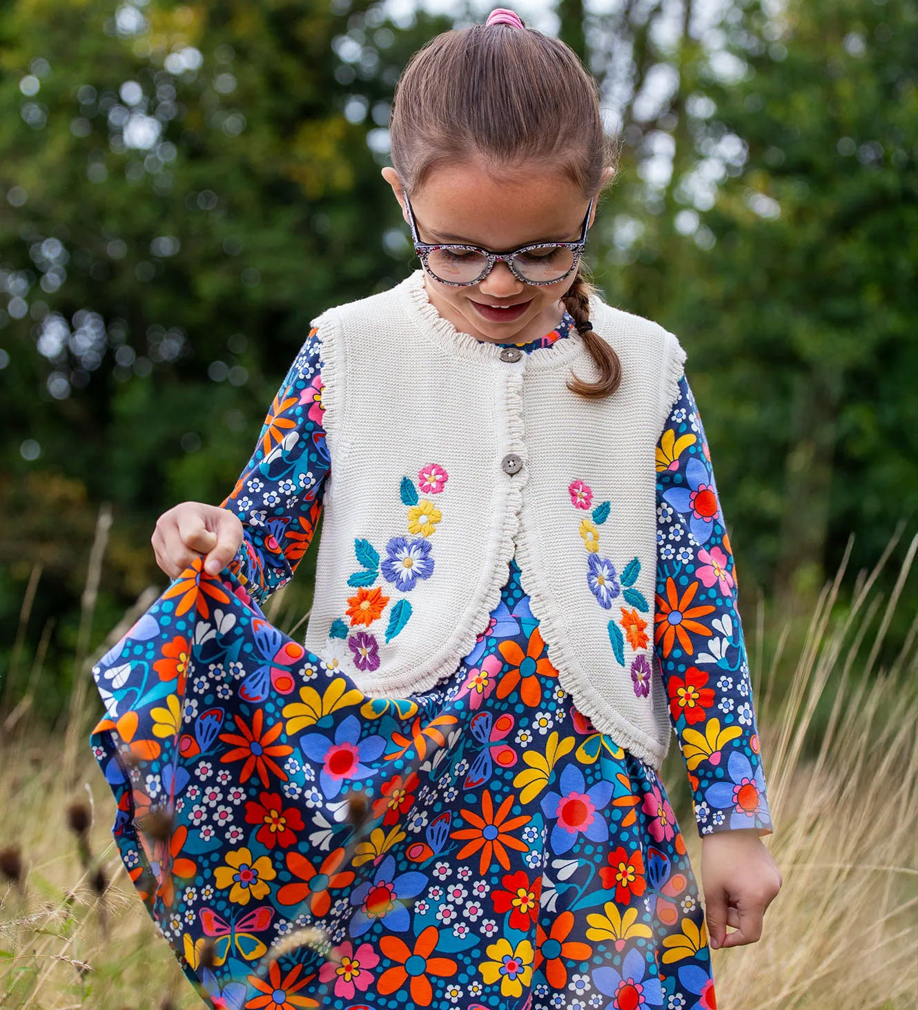 A girl wearing the Frugi Oatmeal Flowers Connie Cotton Knitted Sleeveless Cardigan available at Babipur. This white oatmeal sleeveless cardigan has cute scalloped edges and is decorated with pretty embroidered flowers and leaves on the front.
