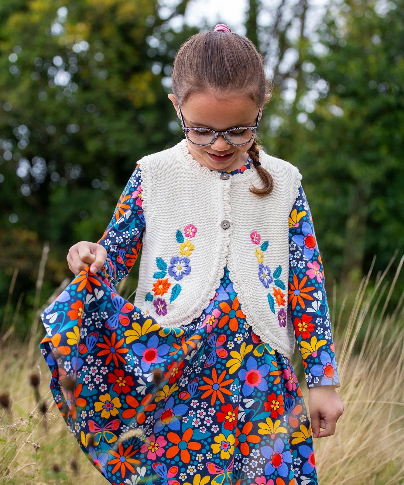 A girl wearing the Frugi Oatmeal Flowers Connie Cotton Knitted Sleeveless Cardigan available at Babipur. This white oatmeal sleeveless cardigan has cute scalloped edges and is decorated with pretty embroidered flowers and leaves on the front.