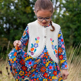 A girl wearing the Frugi Oatmeal Flowers Connie Cotton Knitted Sleeveless Cardigan available at Babipur. This white oatmeal sleeveless cardigan has cute scalloped edges and is decorated with pretty embroidered flowers and leaves on the front.