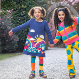 A child wearing the Frugi Kari navy rainbow dress and standing next to another child wearing the Frugi bight rainbow striped cardigan and super dot trousers available at Babipur.