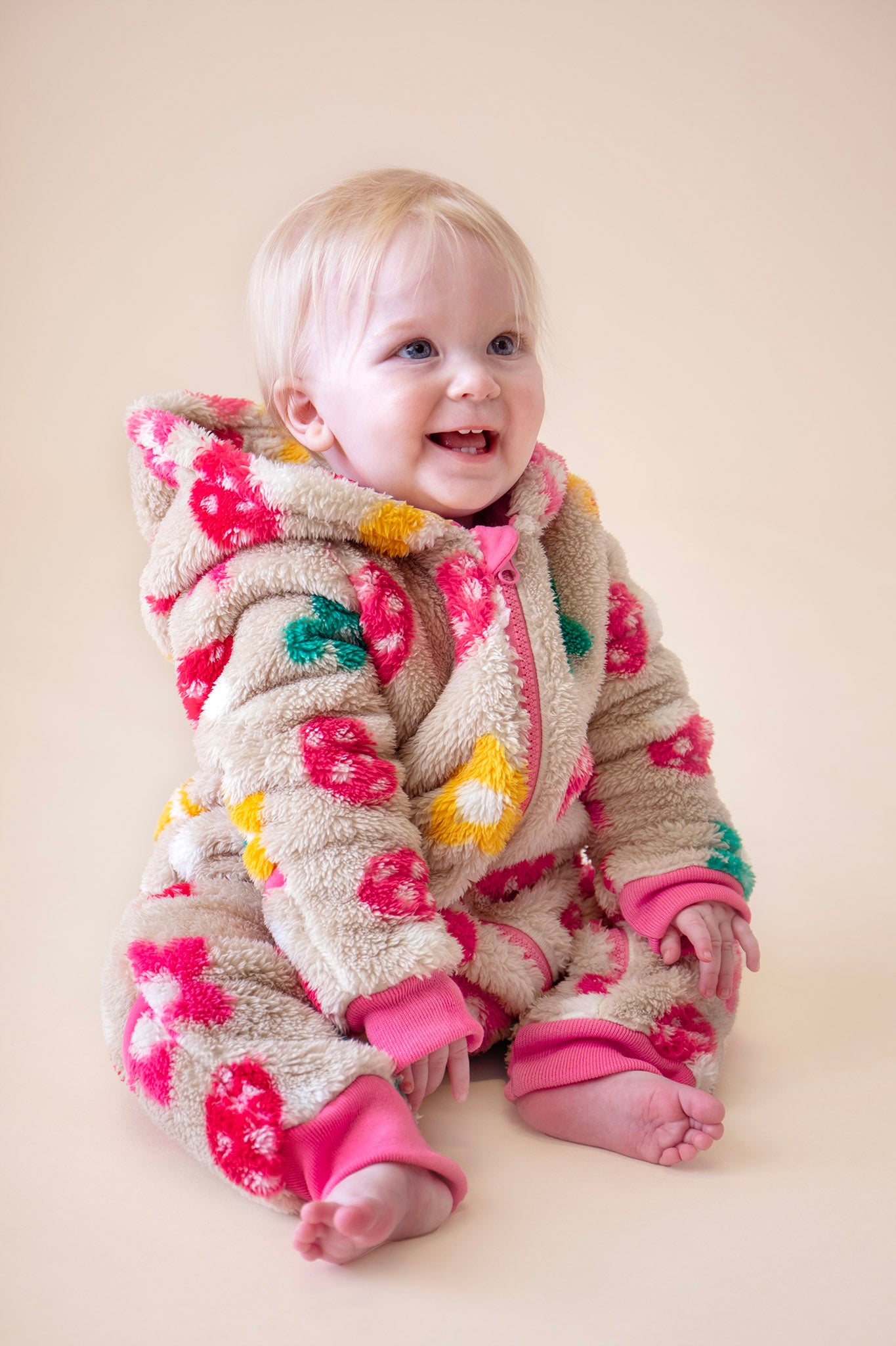 A toddler sitting down and wearing the Frugi kids cosy ted snuggle suit in mushroom field available at Babipur. A thick all in one suit in beige with a cute mushroom and flower design