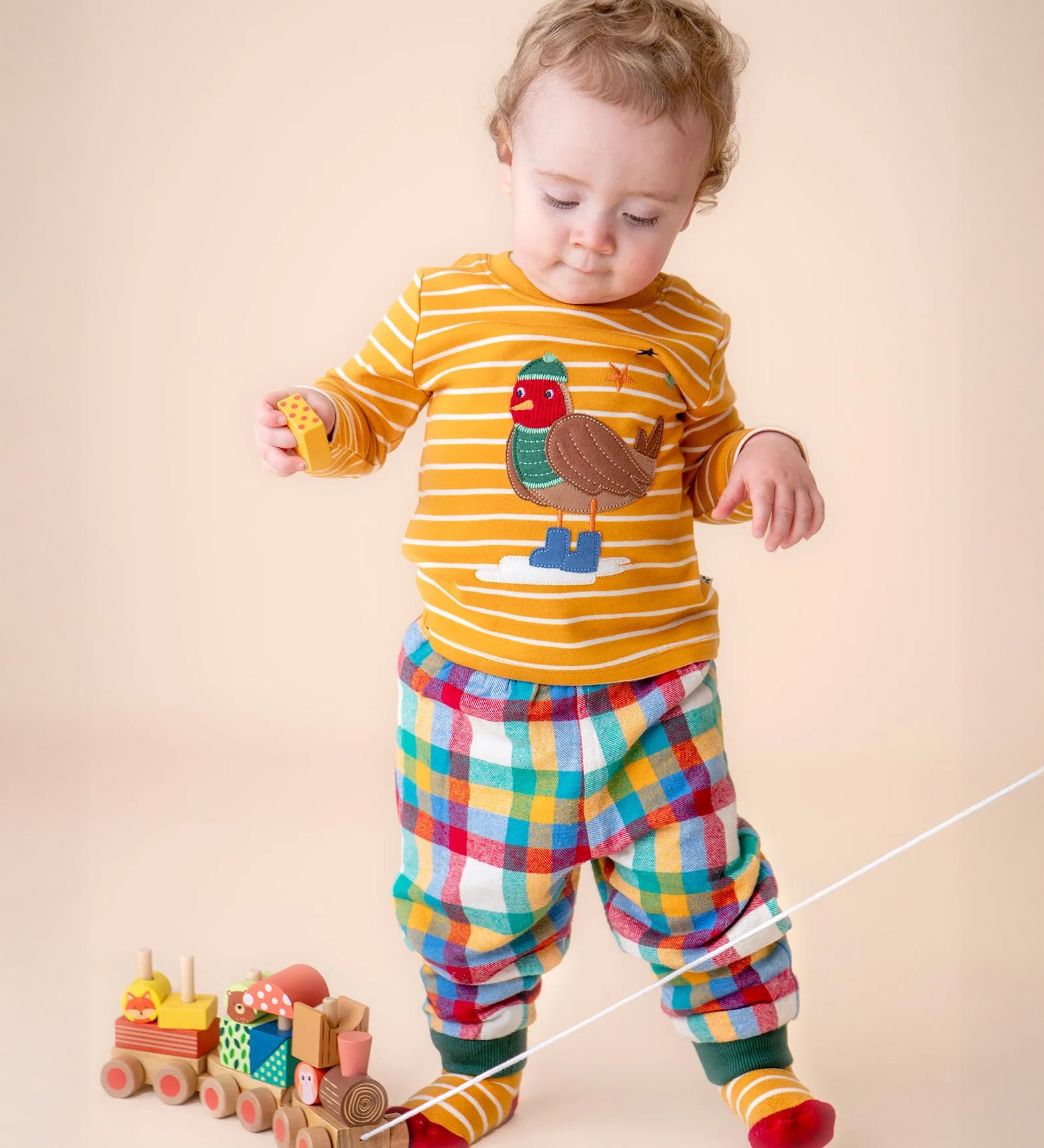 A small child playing with a wooden toy and wearing the Frugi discovery applique top in gold breton/robin and rainbow check bottoms available at Babipur.