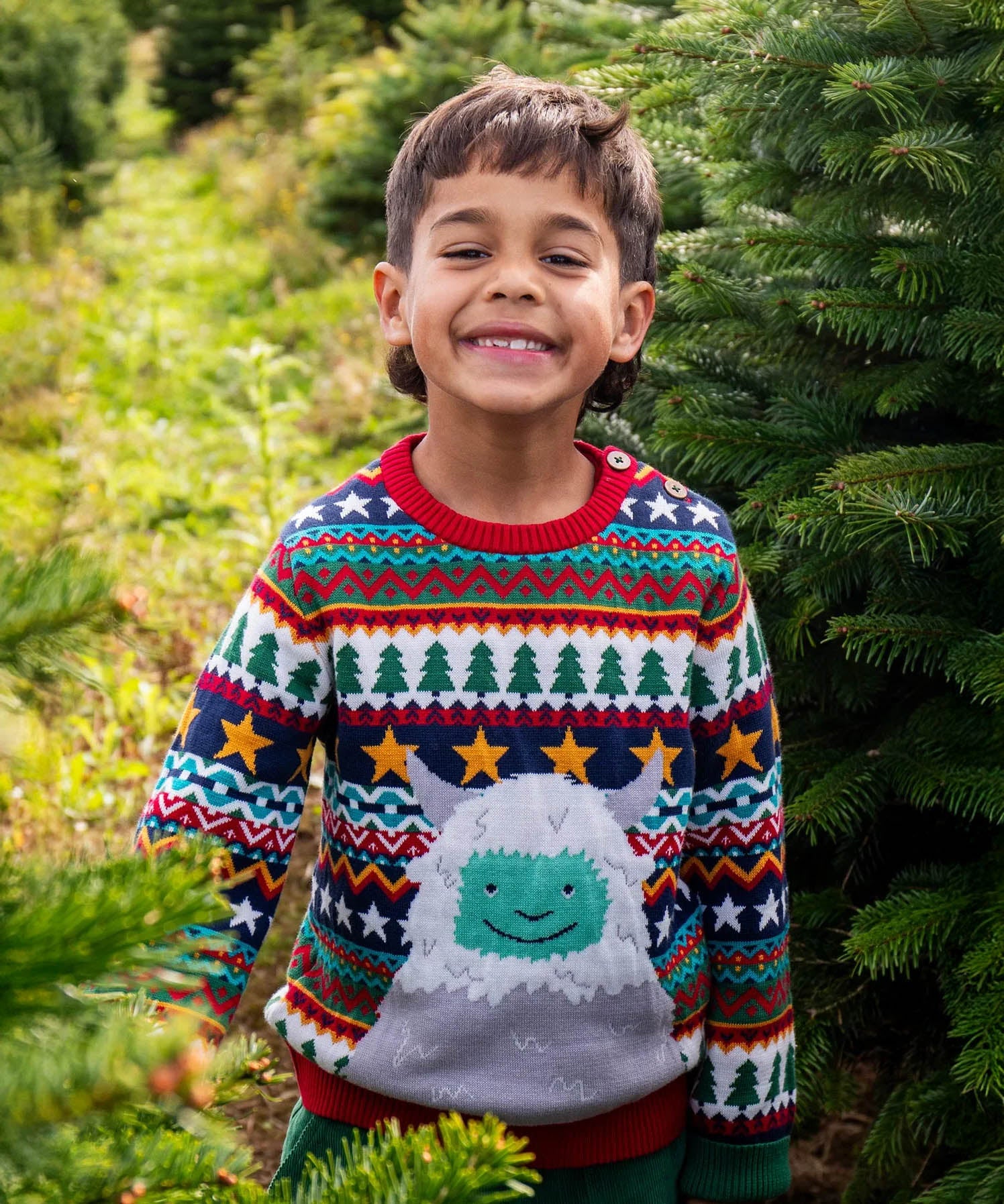 A child standing in a forest and wearinf the Frugi forest fairisle/yeti jumper available at Babipur. A GOTS organic cotton jumper with a cute playful yeti on a festive fairisle design. Features a button fasteners on the shoulder