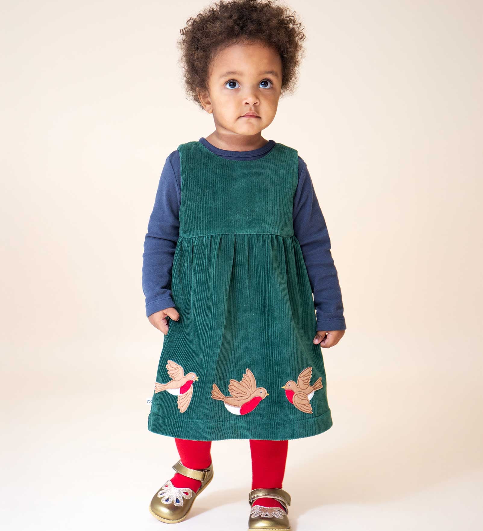 A child wearing the Frugi Rosalie Reversible Baby and Toddler Dress on the green cord side with red tights and gold shoes. One side of this reversible dress features robin redbreast appliques on a Holly green cord with the other side featuring a rainbow coloured check. This dress is a part of a wide range of organic cotton clothing for children available here at Babipur.