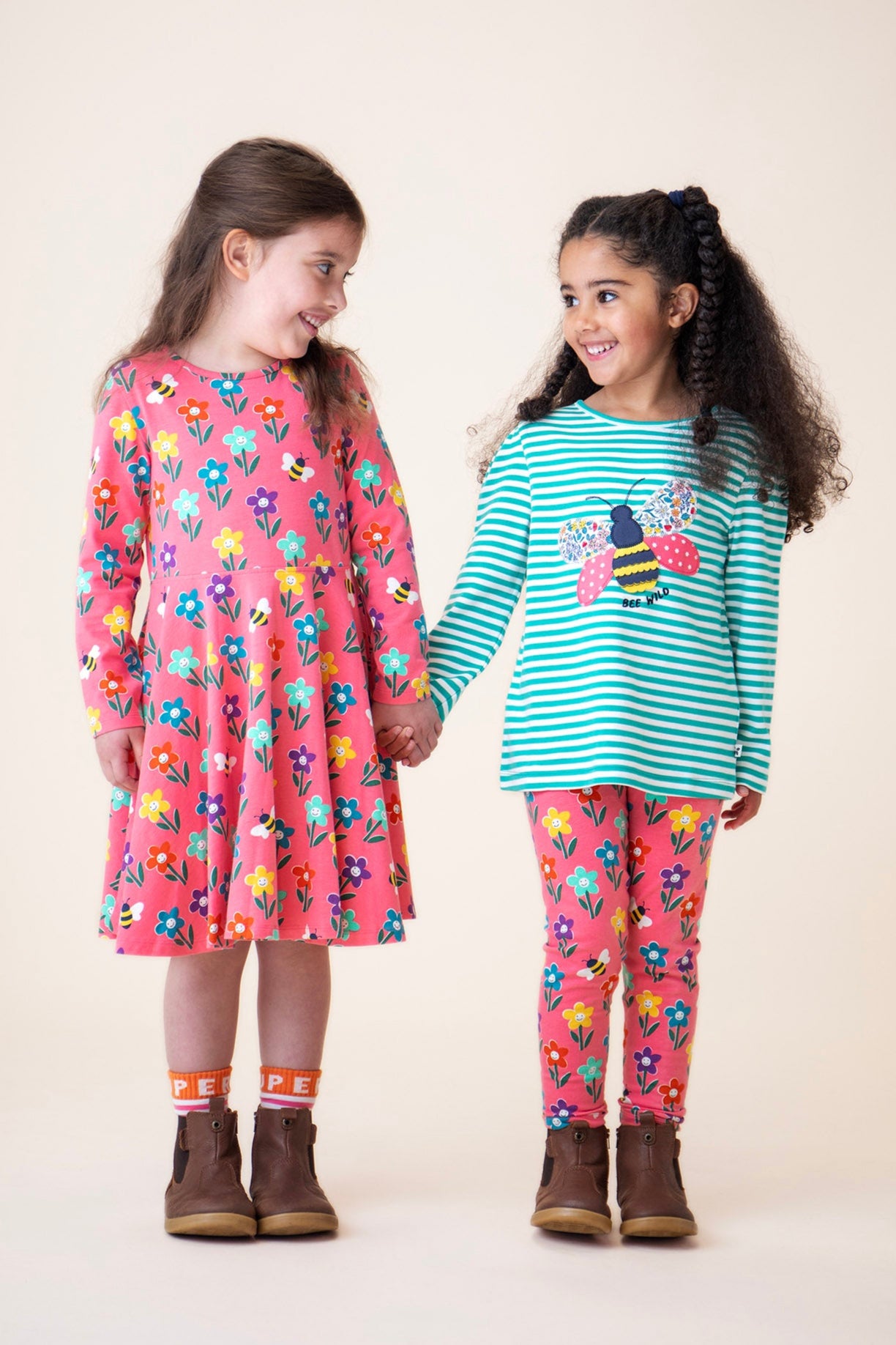A child wearing the Frugi sofia skater dress in tulip/fun flowers and standing next to another child wearing the tulip fun flowers leggings with the Frugi dragon stripe/bee top available at Babipur