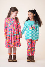A child wearing the Frugi sofia skater dress in tulip/fun flowers and standing next to another child wearing the tulip fun flowers leggings with the Frugi dragon stripe/bee top available at Babipur